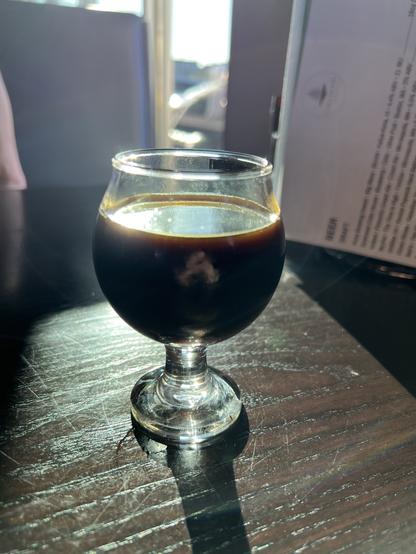 The tiniest of glasses holds an inky dark beer bathed in sunlight.