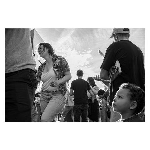 Au Festival de jazz de Montréal, auprès d'un kiosque où un band joue un jazz New Orleans entraînant, curieux et spectateurs dansent. Un enfant les regarde. La photographie est prise en contre-plongée, à hauteur de vue de l'enfant. 
At the Montreal Jazz Festival, people dance beside a bandstand playing lively New Orleans jazz. A child looks on. The photograph is taken from a low angle, at the child's eye level.