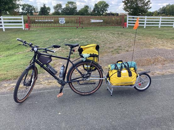 A Priority Bicycles model 600 with a single wheel trailer attached.  The bicycle is black and has a pannier (i.e. Saddle bag) on one side of the rear rack.  Water bottles are in two cages in the frame.  The trailer is a single wheel type attached to the rear axel and is held upright by a double-leg stand and has a large yellow dry bag and tent stowed on it.  In the background is a fence with “old” US Highway 61 signs indicating that Canada is 251 miles in one direcdtion, and New Orleans is 1361 miles in the other direction.