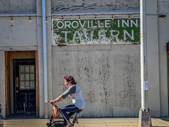An old sign on the wall of the Oroville Inn reads "Oroville Inn Tavern" although the tavern is no longer there. At lower left is a door with a window. A bicyclist rides by in the foreground.
