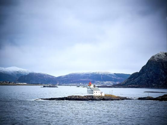 A quaint lighthouse perches on a small rocky outcrop, its white walls stark against the moody blues and grays of the Norwegian seascape. Distant mountains dusted with snow hint at the region's subarctic climate.