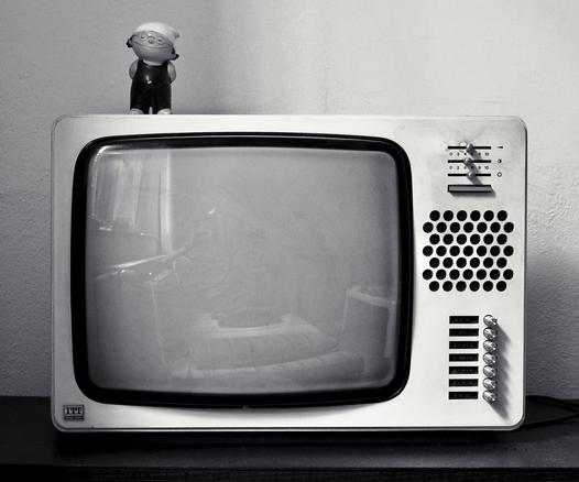 das schwarzweißbild zeigt einen alten schwarzweiß-röhrenfernseher der marke itt. 
der kleine fernseher hat rechts verschiedene bedienelemente für lautstärke, helligkeit und kontrast, die oben zu bedienen sind, sowie sieben drehknöpfe zur programmauswahl unten. dazwischen ist ein grobwabiger bereich, wo der lautsprecher verborgen ist.
der fernseher hat ein weißes gehäuse. links oben auf dem fernseher steht das zdf-mainzelmännchen det.
der hintergrund ist weiße rauhfaserwand. der fernseher steht auf einem dunklen schrank.