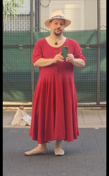 Selbstporträt in spiegelnder Scheibe. Mann mittleren Alters mit dunklen kurzen Haaren und Kinnbart trägt ein rotes Kleid aus Leinen. Das Oberteil hat eine Knopfleiste, kurze Ärmel und einen runden Halsausschnitt ohne Kragen. Der Rockteil ist knöchellang und weit geschnitten, so dass er in Falten hängt. Dazu trage ich einen hellbeigen Strohhut, beige Ballerinas, und eine Halskette mit rundem Holzanhänger.