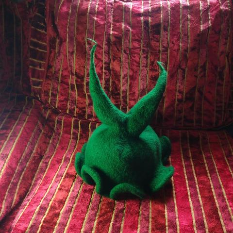 A picture of a green hat with two points and a wavy brim, made of wool. The background is a striped fabric material.