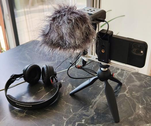 Ein iPhone als live streaming device auf Stativ mit mobilen Rekorder als Mikrofon daneben, über USB Adapter angeschlossen, mit Kopfhörer daneben.