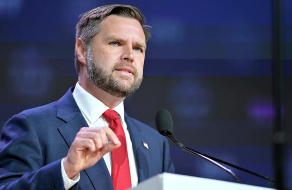 JD Vance behind a lectern is speaking to a recent audience. He wears a dark blue suit, white shirt, and red tie. His right hand is slightly raised in a gesture. The picture is cropped to show only his face and upper body.