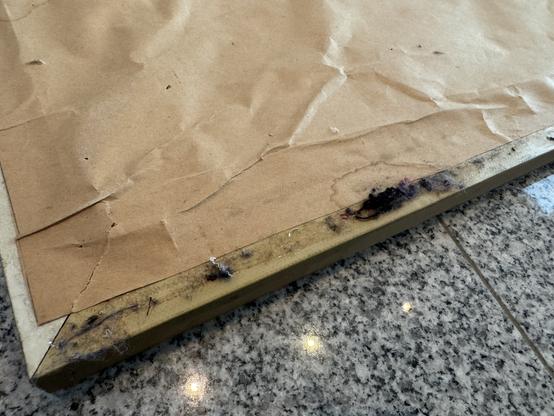 A close up of glue that is still sticky and has picked up all sorts of fiber and natural debris on the edge of a tapestry frame with a poorly applied paper backing. The paper shows the bloom marks from something damp. The fram is resting on a grey granite floor.