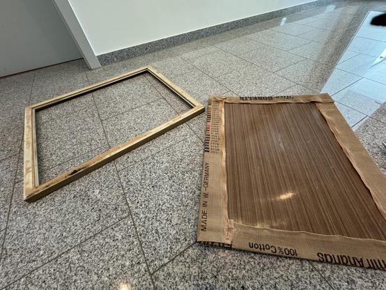 A tapestry out of its frame is face down on a grey granite floor. It has been stretched over a very not-flat sheet of brown faux-wood laminate from the 60’s and stapled in place. At least this one wasn’t trimmed so it will be easy to wash, block and lace in place on a new stretcher frame.
