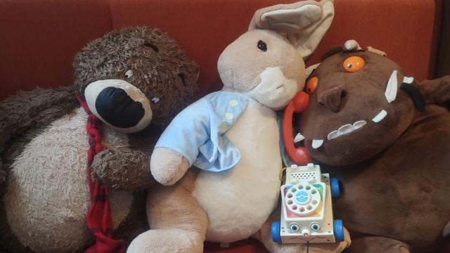 Cuddly Hugless Douglas, Peter Rabbit and Gruffalo plushies slumped together on a seat with a toddler's toy telephone, in the Edinburgh Bookshop's children's room