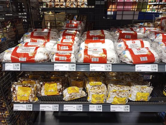 Regale mit verschiedenen Sorten von verpacktem Stollen. Oben liegen weiße, rechteckige Packungen mit rot-weißen Etiketten, die "Edel-Marzipan Stollen", "Butter-Mandel Stollen" und "Christstollen" enthalten. Darunter befinden sich gelbe Verpackungen mit Stollenkonfekt und "Butterstollen Scheiben". Alle Stollen sind in Kunststoffverpackungen eingeschweißt und liegen auf schwarzen Metallgittern. Preise und Produktinformationen sind auf Etiketten an der Regalvorderseite angebracht.