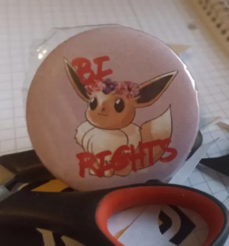 A big round button(the kind you attach to your backpack or clothing), that shows an evoli wearing a ring of pink and purple flowers on the head. Black letters say "BI RIGHTS" in a font, that looks hand written with a felt-tip pen. The background is light pink. The protective plastic wrapping is loose on the upper left edge. The button is secured with the handle of scissors and everything is resting on a college block.