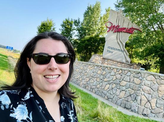 A woman with shoulder-length dark hair and sunglasses on stands on the side of the interstate next to a "Welcome to Minnesota" sign