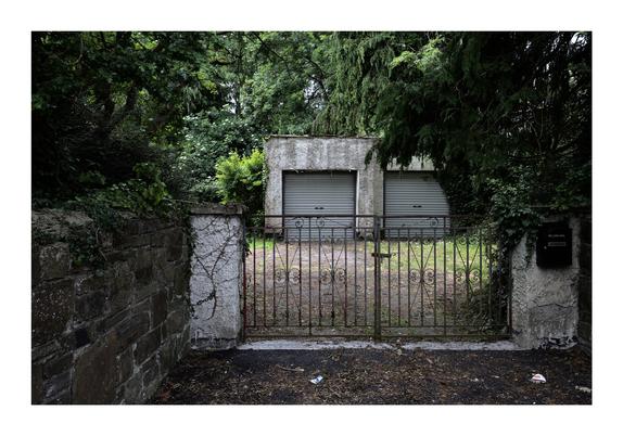 An old, weathered gate stands in front of a small, overgrown driveway leading to a concrete garage with two closed, gray metal doors. The surrounding area is lush with dense green foliage, partially obscuring the garage and the path. The stone walls flanking the gate are covered in ivy and moss, adding to the aged and abandoned feel of the scene.