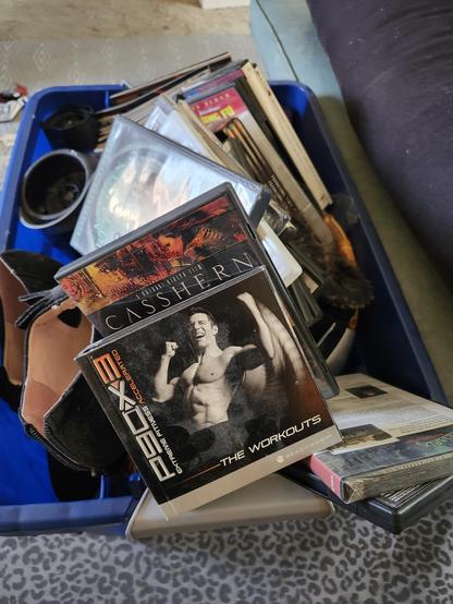 A full bin of clothes, shoes, comics, and dvds, including a p90X3 DVD series (crossfit dude rawr-ing at the camera showing off his completely unnecessary muscles.)
