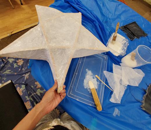 My left hand holding corner of paper mache 3D lantern. Glue & paper supplies on table.