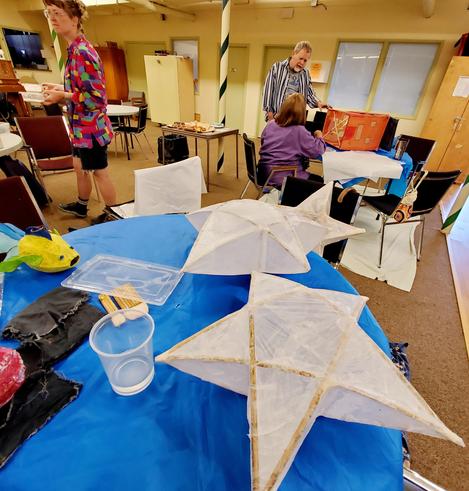 2 large paper mache lanterns, a green & yellow tropical fish paper lantern on table. In background are volunteers working on repairing other paper lanterns.