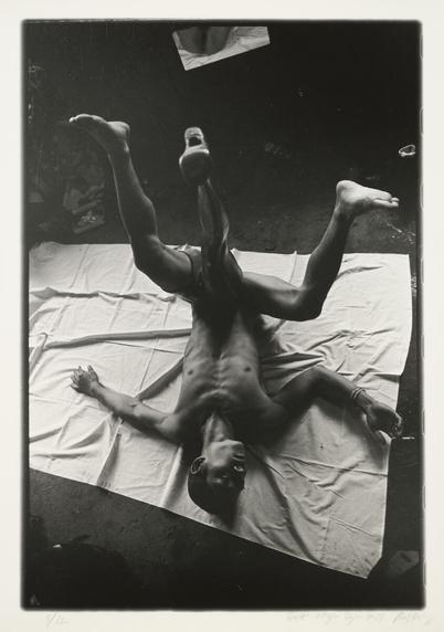 Обнажённый худощавый художник Чжан Хуань лежит на полу на новой белой простыне, подняв свои полусогнутые ноги в воздух в разные стороны. Его руки разведены, в правой он держит сигарету. Снят сверху со стороны головы. К левой ноге художника прикреплён протез цельной ноги, который также задран вверх над полом. Протез находится ровно между двумя настоящими ногами. Основание протеза прикрывает гениталии художника. Сверху видны колени кого-то другого, кто сидит на полу неподалёку. // The nude, skinny artist Zhang Huan lies on the floor on a new white sheet, raising his half-bent legs in the air in different directions. His arms are spread out, in his right he holds a cigarette. Shot from above, from the head side. Attached to the artist’s left leg is a prosthetic leg, which is also raised up above the floor. The prosthesis is located exactly between his two real legs. The base of the prosthesis covers the artist’s genitals. From above, the knees of someone else are visible, who is sitting on the floor nearby. // RongRong, 1993 No. 17 (Zhang Huan), gelatin silver print