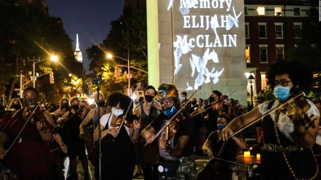 A photograph shows the violin vigil held on June 27, 2020 in downtown Denver. The Colorado capital building is lit in the background, as thousands line the street individual playing their violins. In the center is a sign with illuminated white doves and lettering that reads: "In Memory of Elijah McClain".