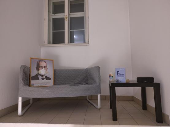 Ein gerahmtes Bild von Alexander v. d. Bellen das auf einem Ikea Knopparp sofa sitzt. Es sieht aus als würde er eine Maske tragen. die Maske ist auf das bild geklebt.