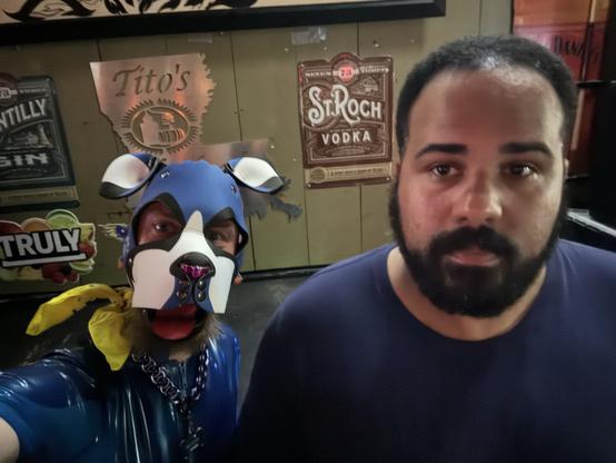 A person wearing a blue dog hood and a yellow bandana poses for a selfie, while another individual with a beard stands nearby. The background features various alcohol-related signage.