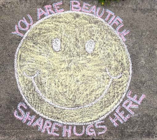 This is a chalk art drawing in front of our house. The main drawing is a large (4' diameter circle, filled in to make a happy face. At the top it reads, "You Are Beautiful." At the bottom it reads, "Share Hugs Here."