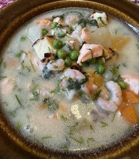 A bowl of soup featuring shrimp, salmon, green peas, and diced vegetables, garnished with fresh herbs in a creamy broth.