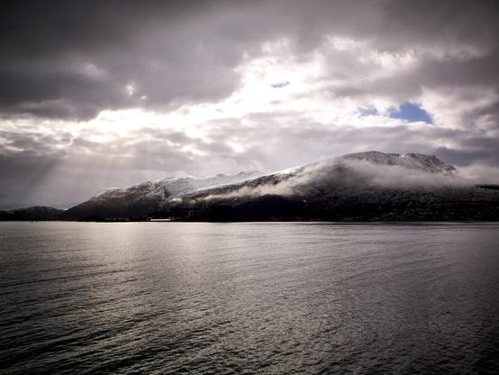 This photo from Norway captures a serene and dramatic landscape. In the foreground, calm waters reflect the muted light of a cloudy sky. The background features a snow-dusted mountain range partially veiled by low-lying mist or fog. Sunbeams break through the dense cloud cover, casting rays of light across the mountain, adding a dynamic and ethereal quality to the scene. The overall atmosphere conveys a sense of tranquility and the raw, untouched beauty typical of Norway's natural landscapes.