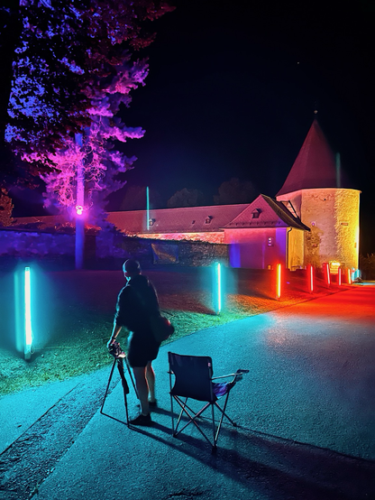 Das Bild zeigt eine nächtliche Szene im Freien, beleuchtet von bunten Lichtern. Im Vordergrund steht eine Person, die eine Kamera und ein Stativ hält, neben einem zusammenklappbaren Campingstuhl. Der Boden ist von blauem Licht erleuchtet, während die Lichter entlang eines Weges in Blau und Rot leuchten. Im Hintergrund ist ein Gebäude mit mittelalterlichem Aussehen zu sehen, das von gelbem Licht angestrahlt wird. Ein Baum auf der linken Seite des Bildes ist von violettem Licht beleuchtet, was der Szene ein surrealistisches Gefühl verleiht.