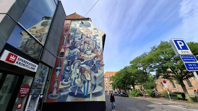 Left, the corner of a financial bank. Right, trees, a church square, a pedestrian, parked cars and bikes. Right in front of us, below bright blue sky: a striking mural on a four-storey gable wall. Painted in earthy tones (plum, rose-pink, dusty blue, browns). It is full of stylised and surreal elements, some reminiscent of Dalí or Picasso. A cactus. A blue avocado with a huge eyeball balanced on it. Fruit. Insects. Central: a woman at a café table with coffee and food on it. She is wearing a hat whose brim is a plate of fruit that has a tre growing out of it. Appropriately for a city where this happens often, a seagull is stealing food from her plate.  The artist said: ‘the seagull stealing food symbolizes the need to remain calm and composed amidst life's unexpected challenges. This scene serves as a reminder to stay vigilant about our physical health, including the importance of a healthy diet.’