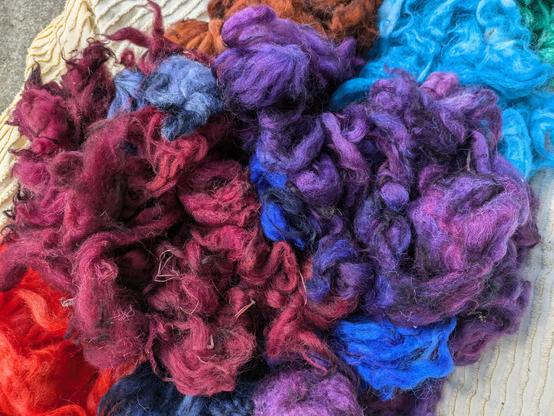 Gefärbte Wollflocken in rot, violett, blau und grün liegen als Haufen auf einem gelben Tuch