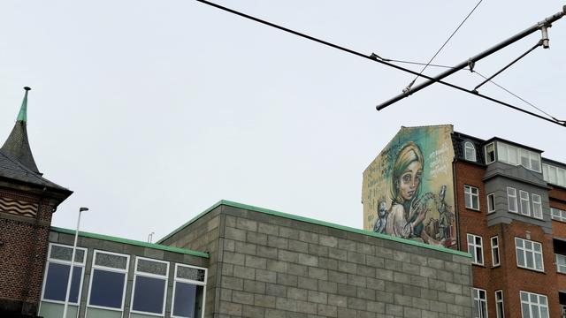 On the high upper part of a gable wall between other historic old buildings, framed by tram cables and a turret, a mural in muted, elegant blue and brown tones. Not all visible, because too inaccessible to photograph fully, it depicts a wide-eyed young woman in trousers sitting on a floor, head turned to look expressionlessly at us. Behind her, a duckling perched on a stack of books. In front of her, Thumbelina, wearing a thimble, head also turned to look at us. Between them, piles of matchboxes and a ladder bridge that they are making out of the matchsticks. The duckling is wearing a crown of matchsticks. Thumbelina is wearing matchstick wings. Around the trio, the words: ‘Just imagine if they let us in … we really could be anything.’