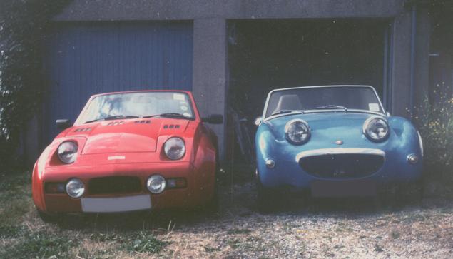 Front view of two sports cars parked in front of a double garage: on the left a red Midas Gold Convertible from 1989, on the right a blue Austin Healey Sprite from around 1959. Both are open-topped and have characteristic frog-eye headlights, and both look like they're smiling