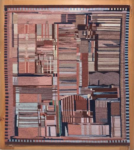 A Navajo rug with a complex pattern with muted reds, pinks and blues. The pattern consists of various vertical and horizontal rectangles with stripes. Around the border are small alternating black and colored rectangles. The weaving is mounted in a wooden frame and hanging on the museum wall.