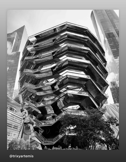 Black and white photo of The Vessel. The vessel building is at the center of the shot and a few other skyscrapers can be seen around the edges of the shot. It is a structure and visitor attraction built as part of the Hudson Yards Redevelopment Project in Manhattan, New York City, New York. Plans  by the British designer Thomas Heatherwick, it has an elaborate honeycomb-like structure rises 16 stories high.