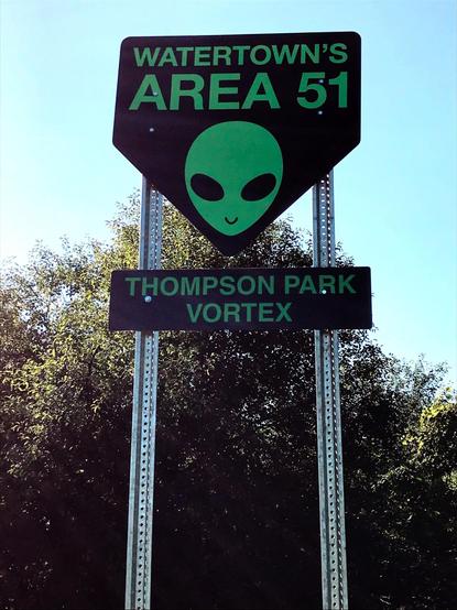 A pair of signs, one over the other, each dark but with pale green lettering. The top sign is shaped like a downward-pointing pentagon and says "Watertown's Area 51" over a picture of a pale green alien head. The bottom sign is rectangular and says "Thompson Park Vortex." Behind the sign are trees and blue sky.