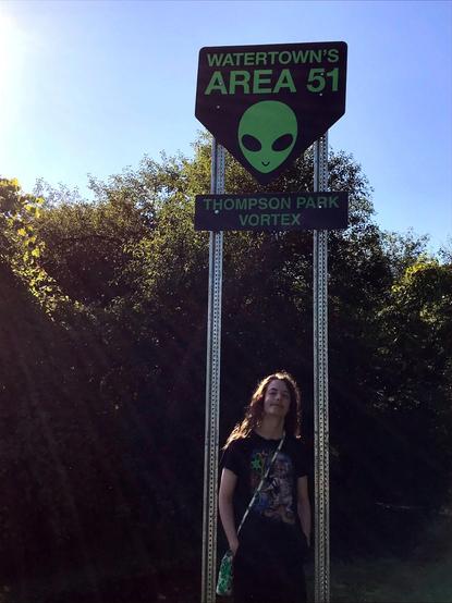 The same signs* as in the first picture, but a long-haired teenaged human stands beneath, hands in pockets. The human wears a cross-body crocheted water bottle holder and a black T shirt which depicts cats riding fidget spinners through outer space.
(*The signs are a pair of signs, one over the other, each dark but with pale green lettering. The top sign is shaped like a downward-pointing pentagon and says "Watertown's Area 51" over a picture of a pale green alien head. The bottom sign is rectangular and says "Thompson Park Vortex." Behind the sign are trees and blue sky.)