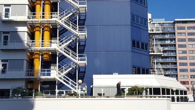 Man in black clothes on the phone on the rooftop terrace of a white building with potted plants and a windowed room. The building is in front of a grey modular one with white fire escape stairs wrapping around large yellow industrial pipes running down the side of the building. Which are in front of the blue rectangular with four windows on the far side modular part of the building. They tower over the terrace, as do the buildings further back in the background.