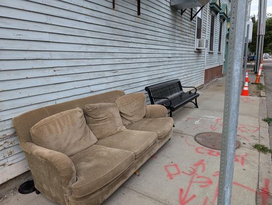 Photo of an overstuffed, velour couch on the sidewalk next to an  austere, metal bench.