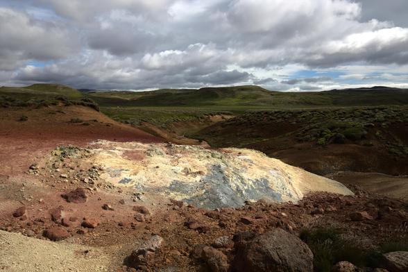 A barren, rocky landscape under a partly cloudy sky with patches of sunlight illuminating the ground. The terrain is a mix of red and brown earth, with pale and dark mineral deposits scattered across the surface. In the distance, green hills roll under the cloudy sky, creating a stark contrast with the arid foreground.