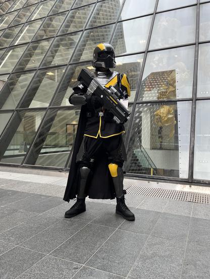A full shot of a figure wearing a costume of dark metallic gray and yellow armor, helmet, and black cape from the game Helldivers 2, holding a prop rifle, standing in front of an array of angled glass windows in downtown Atlanta.