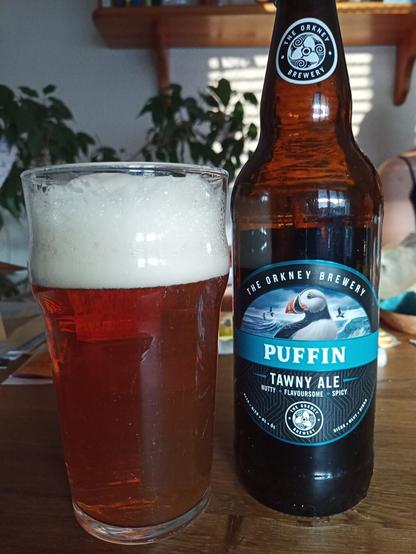 Auf einem Tisch steht ein Glas mit rötlich klarem Bier gefüllt.
Daneben eine Bierflasche aus Schottland mit einem Puffin als Logo auf dem Etikett