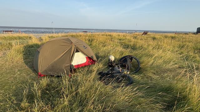 Eine Campingszene mit einem Zelt, das in hohem Gras in der Nähe eines Gewässers aufgestellt ist. Ein Fahrrad liegt neben dem Zelt auf dem Boden.