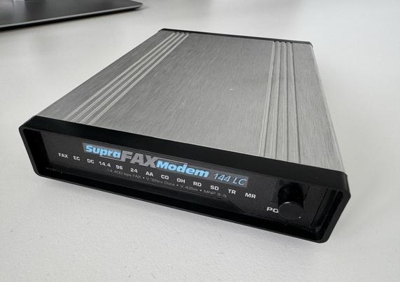 Schräge Frontalansicht eines Supra FAX Modem 144LC im Metallgehäuse mit schwarzer Plastikblende und LEDs für FAX, EC, DC, 14.4, 9.6, 2.4 AA, CD, OH, RD, SD, TR, MR und ein Einschaltknopf.