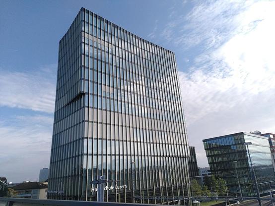 Photo of a tall modern building under a blue cloudy sky. It is the Zug-Rotkreuz campus of the computer science faculty of the University of Applied Sciences of Lucerne.