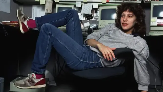 Susan lays back in an office chair, wearing casual clothes, behind her the 80's Apple computers she helped design the operating systems for.