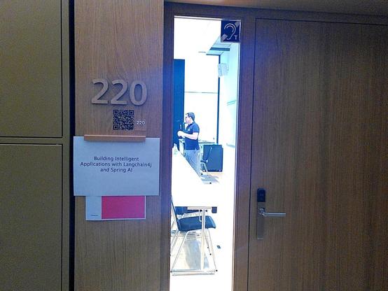 A peek inside the classroom, a presenter is speaking, on the door numbered 220 the text is written "Building intelligent Applications with Langchain4j and Spring AI"