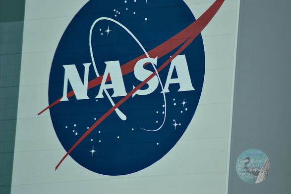 A detailed photograph of the NASA logo on the side of a building.
The logo is a blue circle with the acronym NASA written in plain text.
Stars are scattered through the blue circle. A white ring orbits the letters. A red V-shape with its point on the upper right, meant to imply a vector in space.