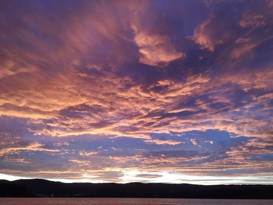Un atardecer con un gran cielo nuboso sobre el mar y las montañas. La luz refleja en las nubes intensos colores púrpuras y salmón mientras todo se cubre de penumbra.