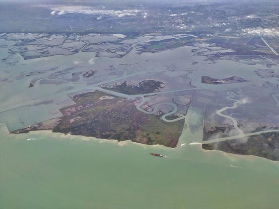 Vista aérea de los alrededores de Venecia. En ella se aprecian las verdosas aguas inundando la tierra fragmentada, y donde asoman serpenteantes ríos. Un barco mercante navega en la parte inferior de la fotografía, pudiendo comparar el inmenso tamaño del área.