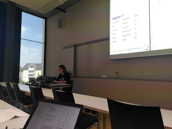 A person at a desk in front of a window and slide presentation. A laptop with code is visible in the foreground.